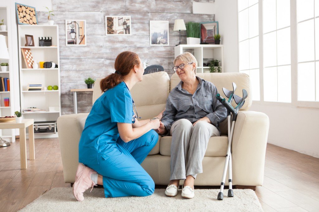 Visiting carer kneeling beside an elderly woman on the sofa, holding her hand with warmth and compassion